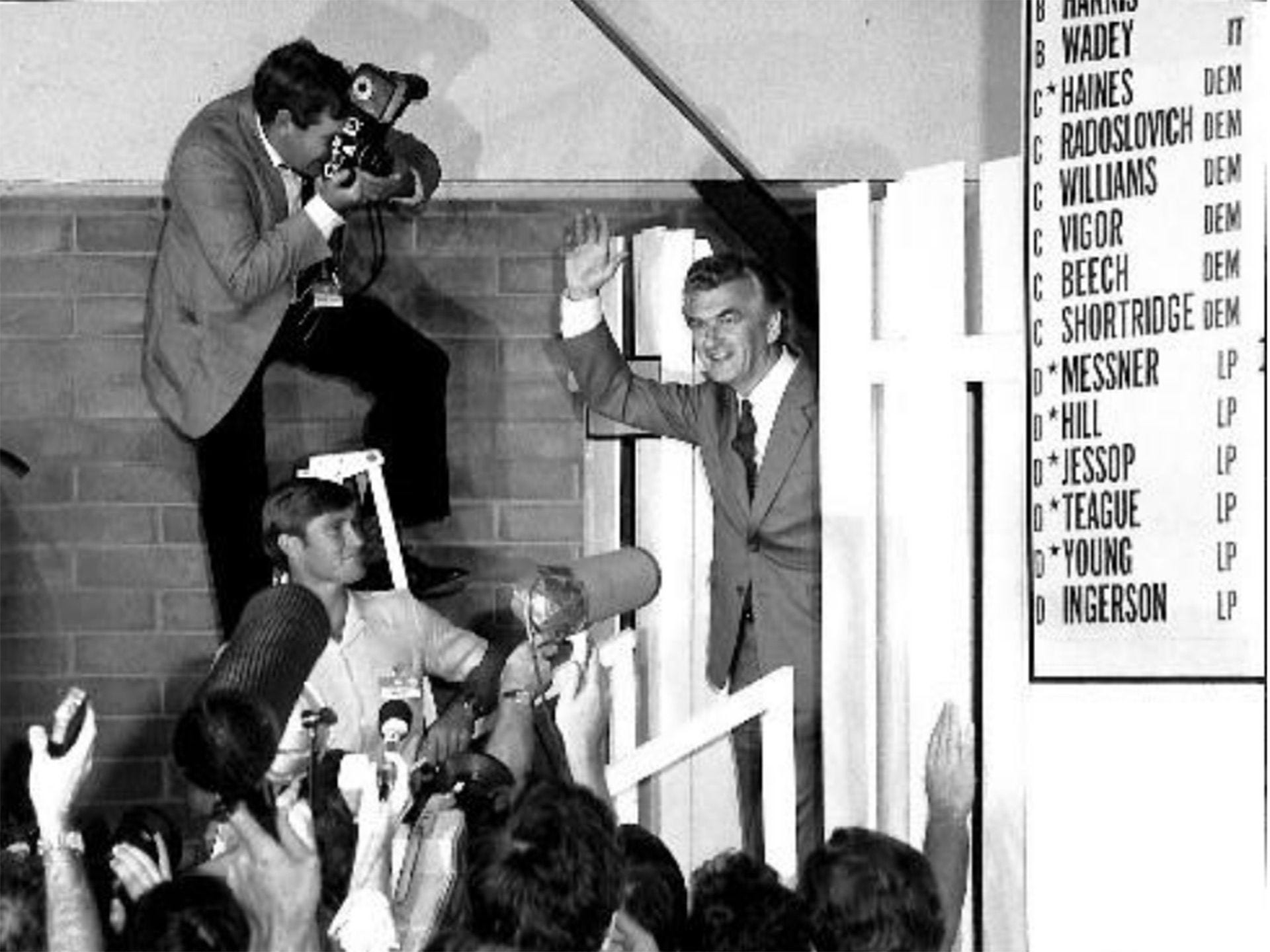 Black and white photo of Bob Hawke waving to a crowd with some media present taking photos of him.