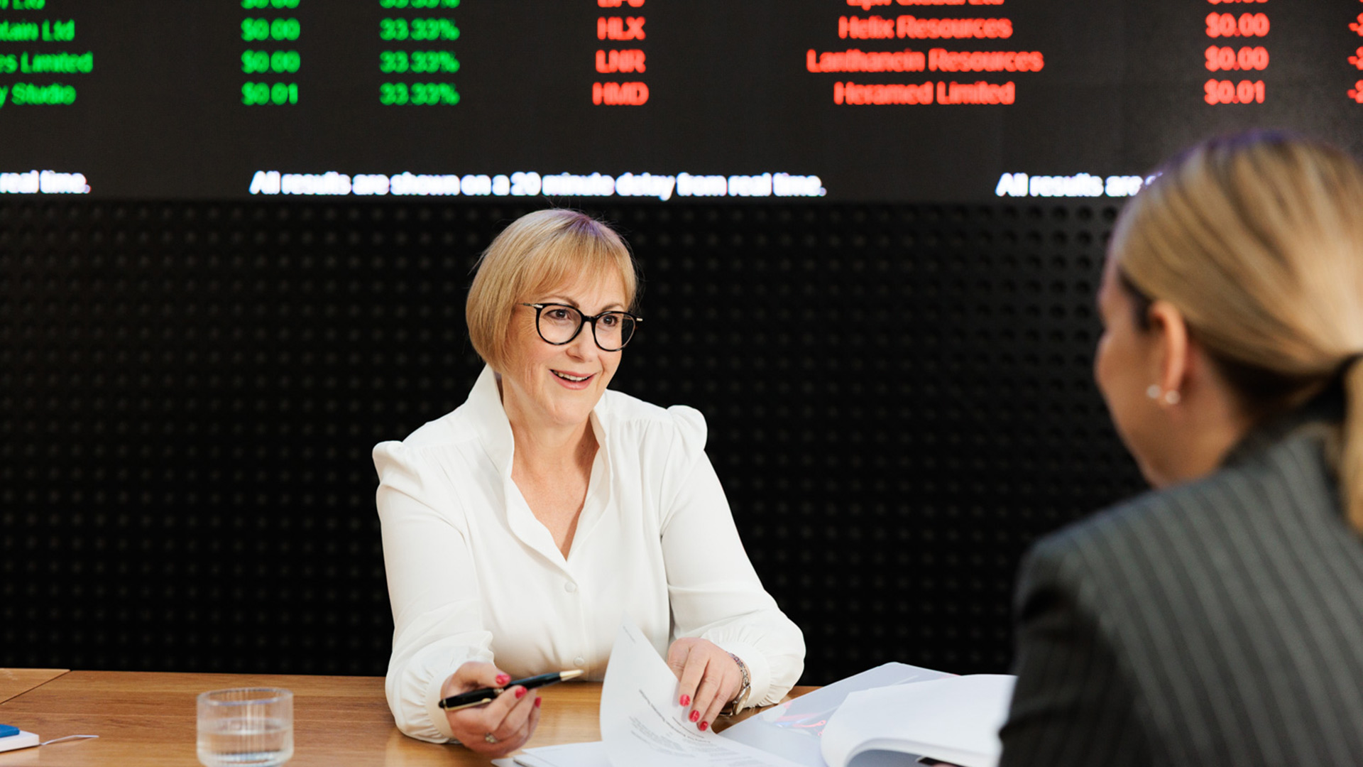 Two business people talking with the stock markets shown behind them.