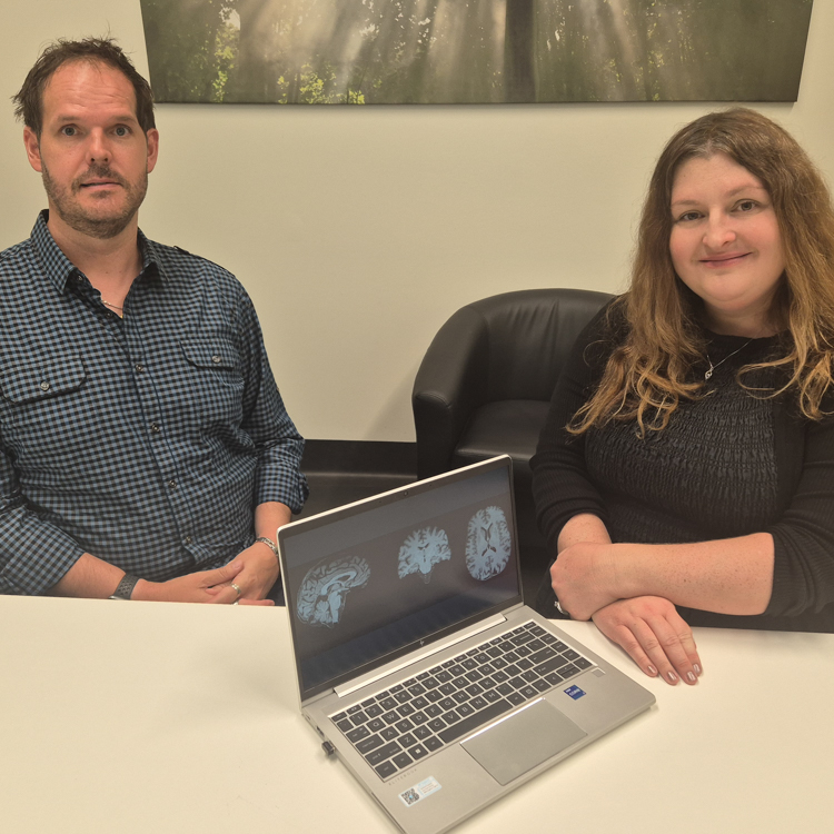 A photo of Associate Professor Lyndsey Collins-Praino sitting at a desk next to trial participant. A laptop displaying photos of brain scans is on the table and facing the camera.